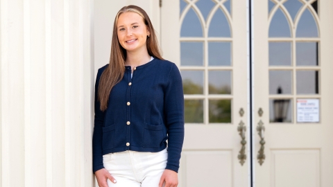 Madelyn Hudak wears a blue top and white pants and leans slightly against a white column outside a building on campus.