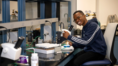 Monwabisi Qeki sits in a chair in a lab setting and smiles as he adjusts a microcope.