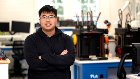 Richard Chi stands in a makerspace and smiles while wearing glasses and a navy hoodie.