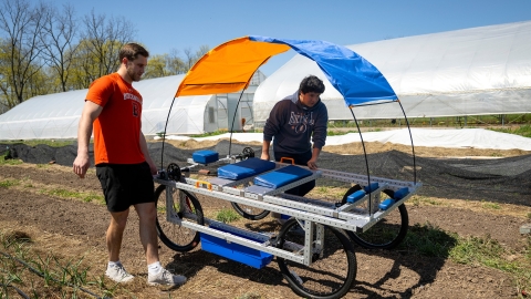 Two students walk along either side of a cart-like machine on four wheels with an orange and blue sunshade and a hoophouse in the background at a farm.