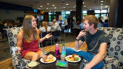 Two students sit in The Bison on colorful chairs and talk while eating food.