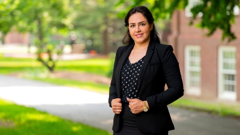 Vaji Farhadi wears a black suit and smiles while standing outside on campus with green trees and sunshine behind her.