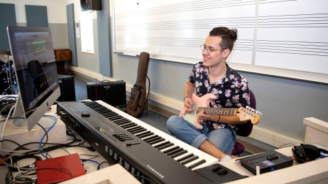 Ian Herdt plays a guitar in front of a musical keyboard and computer