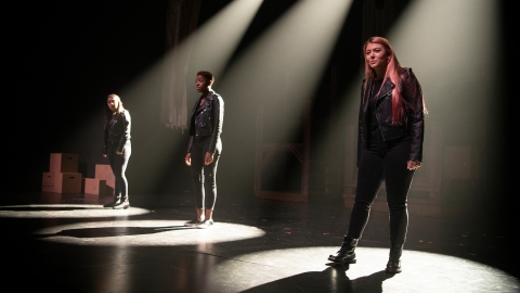 Three actresses stand on stage under spotlights.