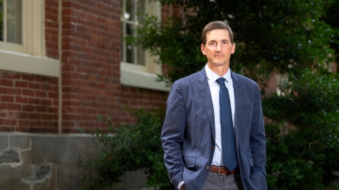 Professor Matt Bailey, in a blue jacket and tie, stands beside a brick building on Bucknell's campus