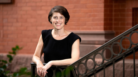 Reggie Gazes wears a black sleeveless shirt and stands against the railing of a staircase outdoors on campus with a red brick building behind her.