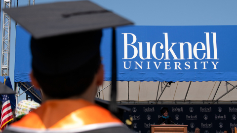 Back of student at commencement wearing a cap and orange sash. Blue banner that says Bucknell University in the background. 