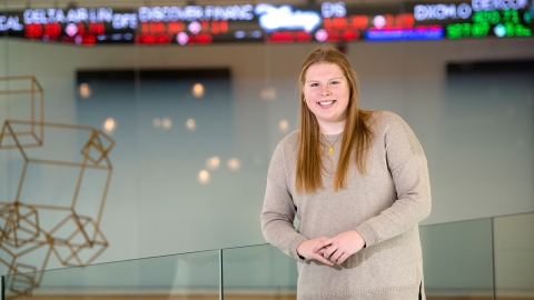 A student stands inside a building with a high-tech ticker behind her, displaying current stock prices. 