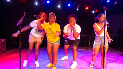 Four students sing on a stage flooded with pink light.