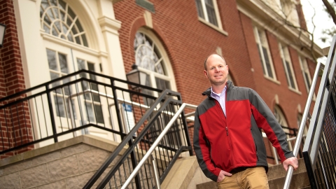 Eric Kennedy wears a red and gray fleece jacket and stands and smiles on the steps outside a building.