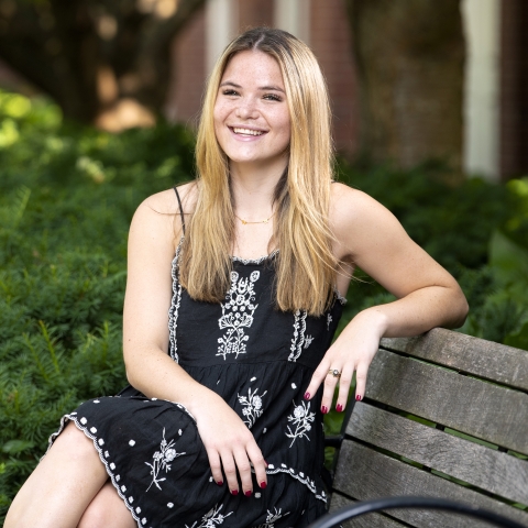 Student in a black dress smiling on a park bench surrounded by greenery