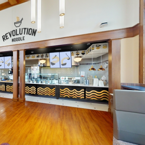 An artist rendering of an Asian takeout restaurant in MacDonald Commons. A sign stating Revolution Noodle appears above a serving counter with instant noodle patterns along the front. A lounge area with leather chairs is in the foreground.