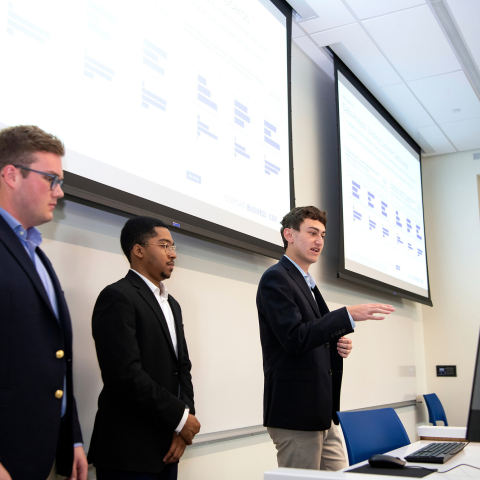 Three students in dark suits give a presentation in front of a whiteboard and projector.
