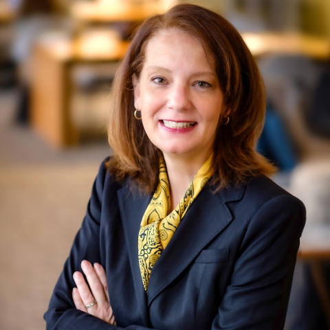 Portrait of Michelle Roehm from the waist up. She is standing with her arms crossed, wearing a blue sport jacket and yellow scarf