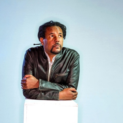 Portrait of author Colson Whitehead leaning forward against a white box, staring upward and off camera to the right.
