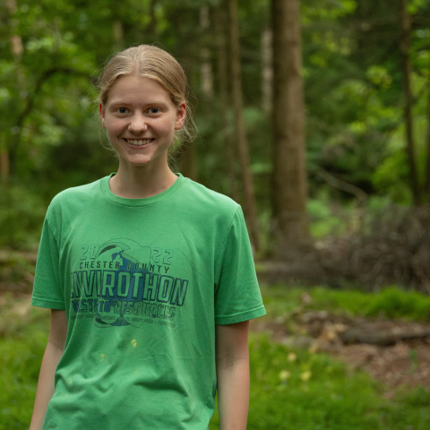 Susan Deering '26 smiles while wearing a green T-shirt in a wooded environment.