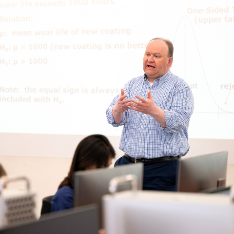 Joe Wilck teaches in a classroom, with a screen filled with equations behind him and students facing him.