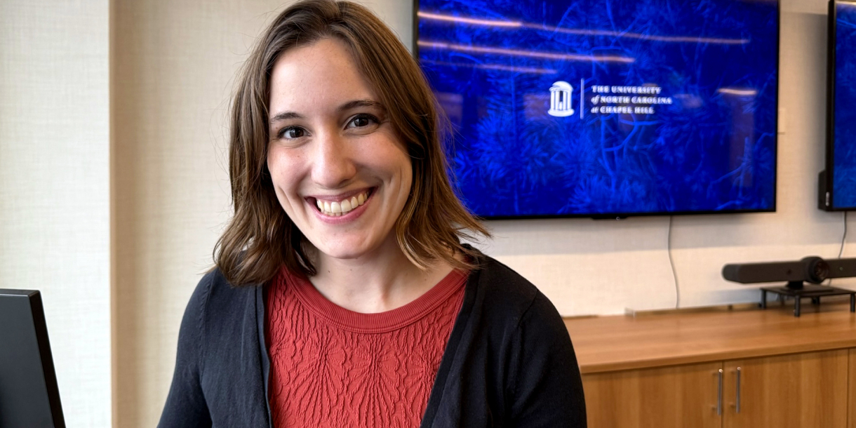 Juliet King smiles in a room where a large screen is on the wall behind her with the University of North Carolina's logo