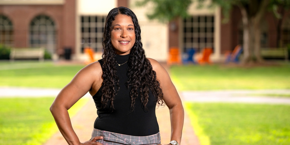 Jasmine Jones &#039;25 stands on campus on a brick walkway with green grass on both sides of it, with her hands on her hips and smiling.