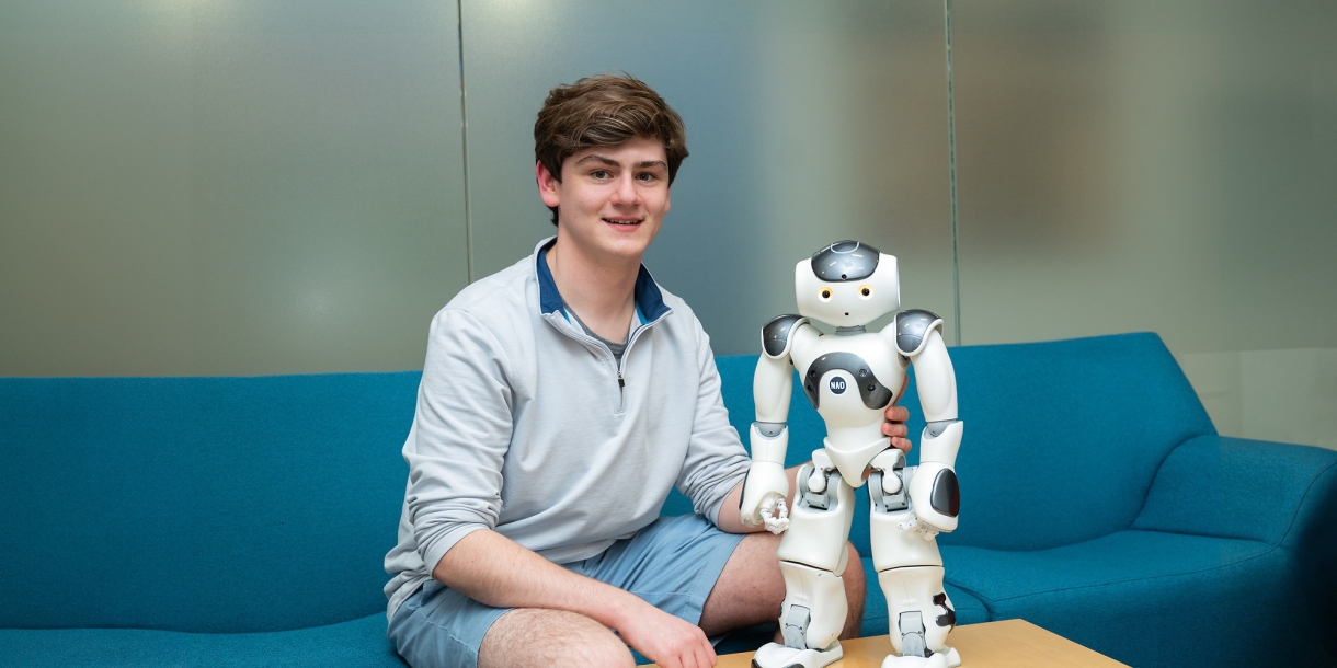 Sean O'Connor sits on a blue couch smiling next to a robot.