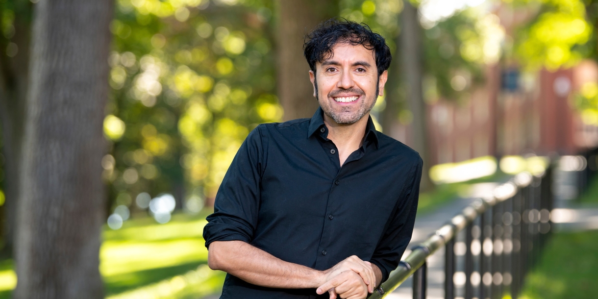 Portrait of Eddy Lopez leaning on a railing outdoors in a wooded area.