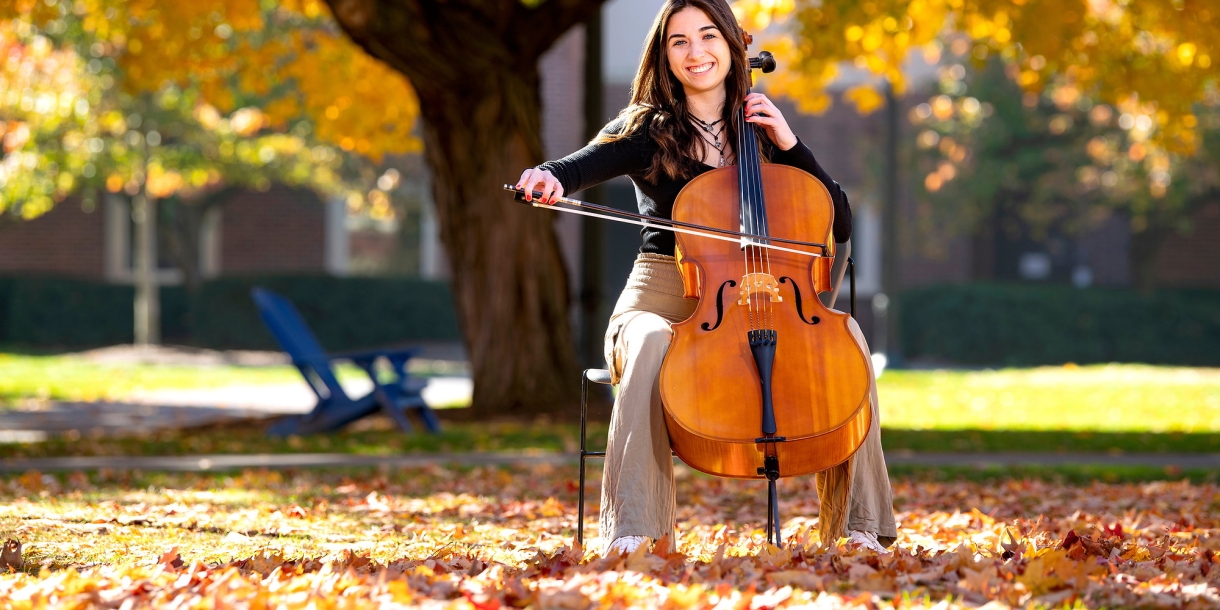 Elle Chrampanis sits on a chair and plays the cello while sitting outside under a tree with golden leaves that hang from the branches and also sit on the ground around her..
