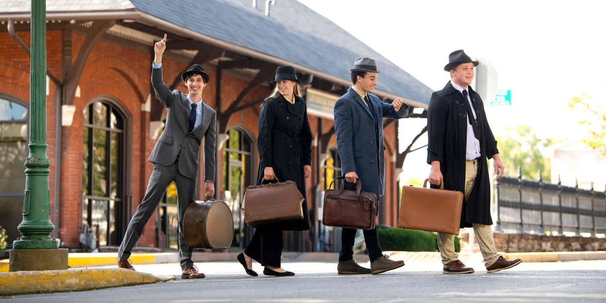 Four people dressed as salesmen cross an intersection in front of a trainstation.