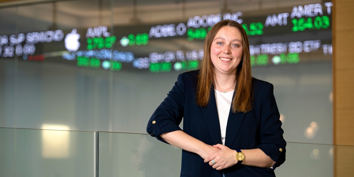 Caitlin Yant &#039;24 stands and smiles in the Moriatry Investment Center in Holmes Hall with a stock ticker behind her.