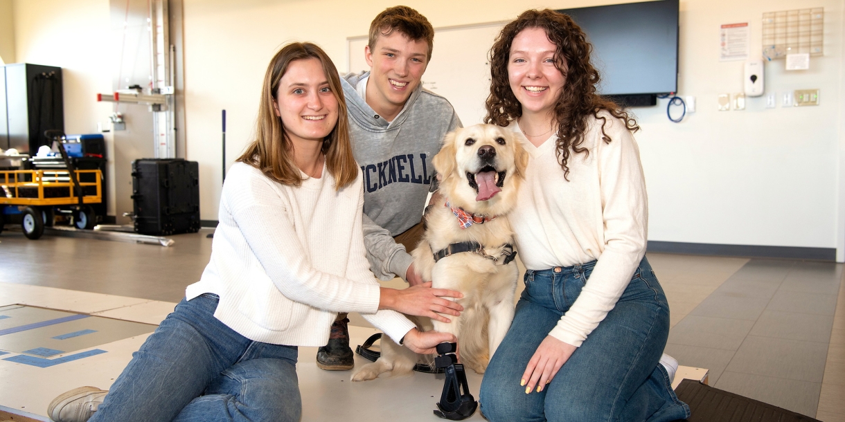 Three students sit and smile with a golden retriever on the ground inside a work space. The dog wears a prosthetic on its front right leg.
