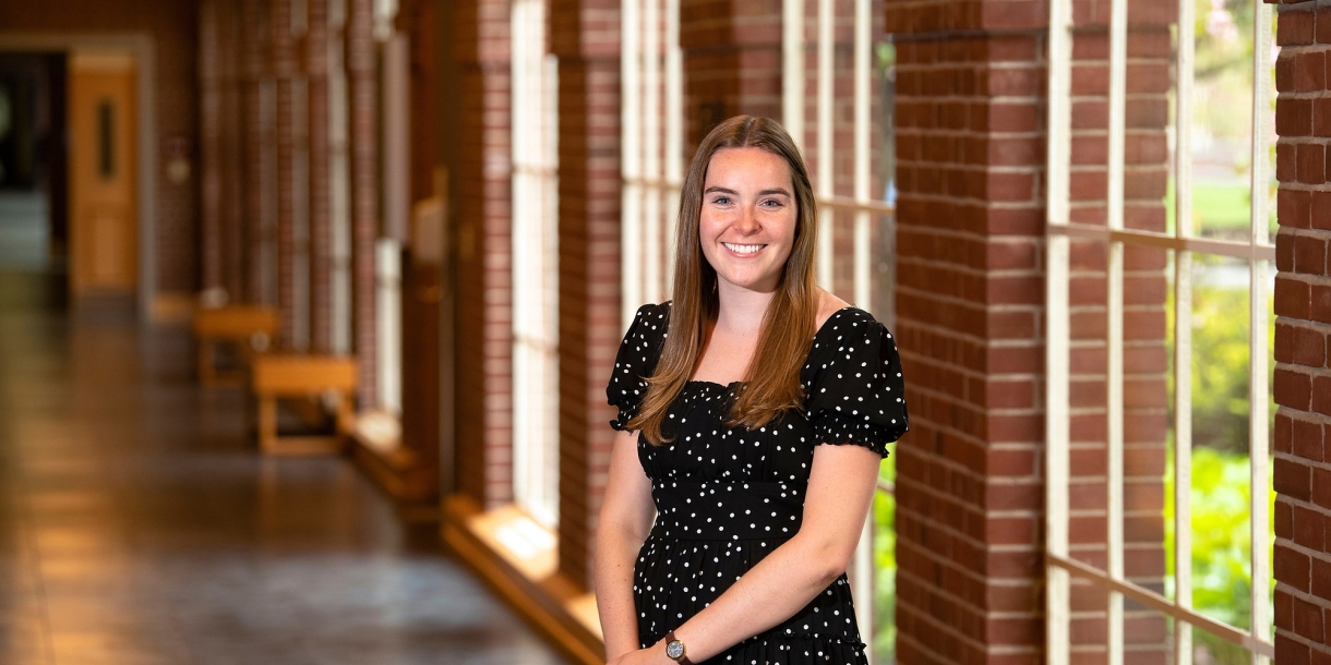 Kendall Robertson wears a black dress and smiles while standing in a hallway with brick columns and windows behind her.