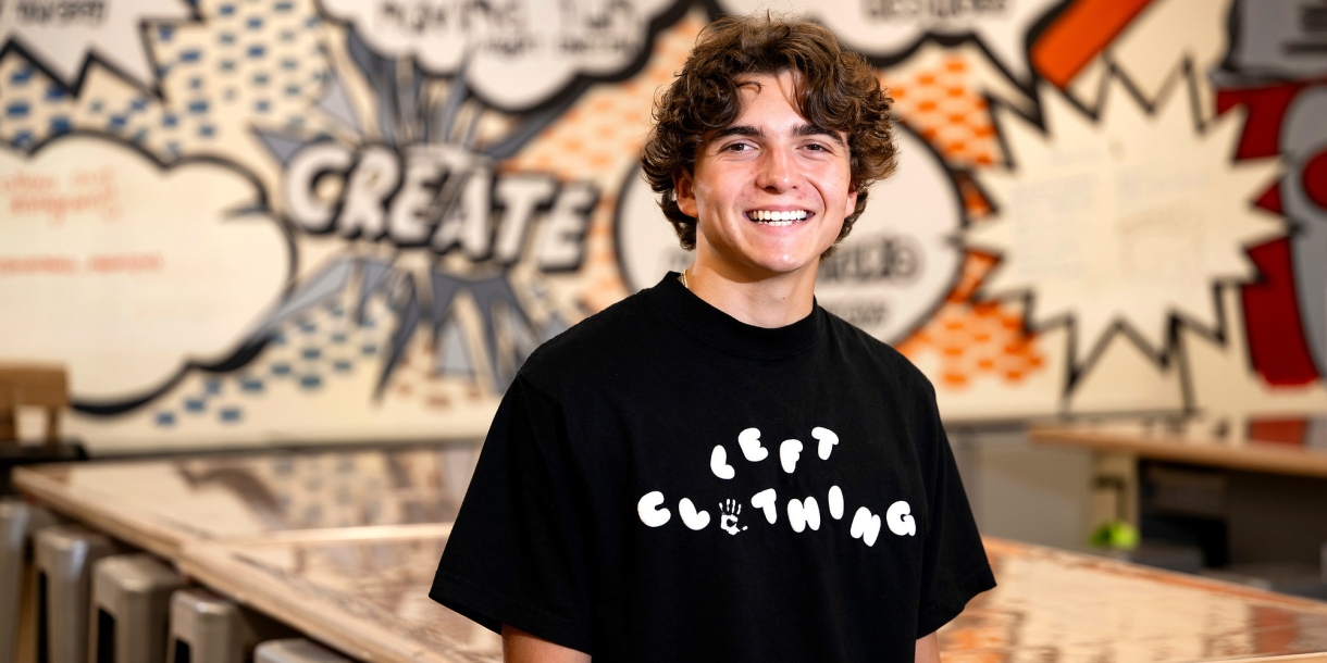 Mikey Brandt wears a black Tshirt with a LEFT Clothing logo printed across his chest. He is in a Makerspace with a graffiti-style wall design behind him.
