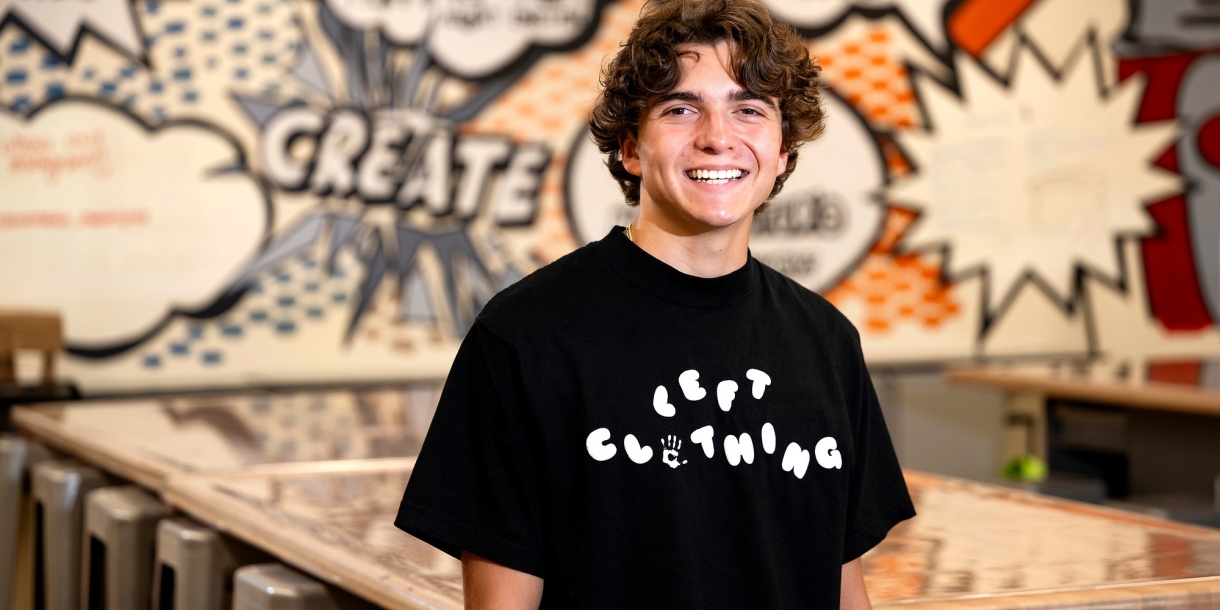 Mikey Brandt wears a black Tshirt with a LEFT Clothing logo printed across his chest. He is in a Makerspace with a graffiti-style wall design behind him.