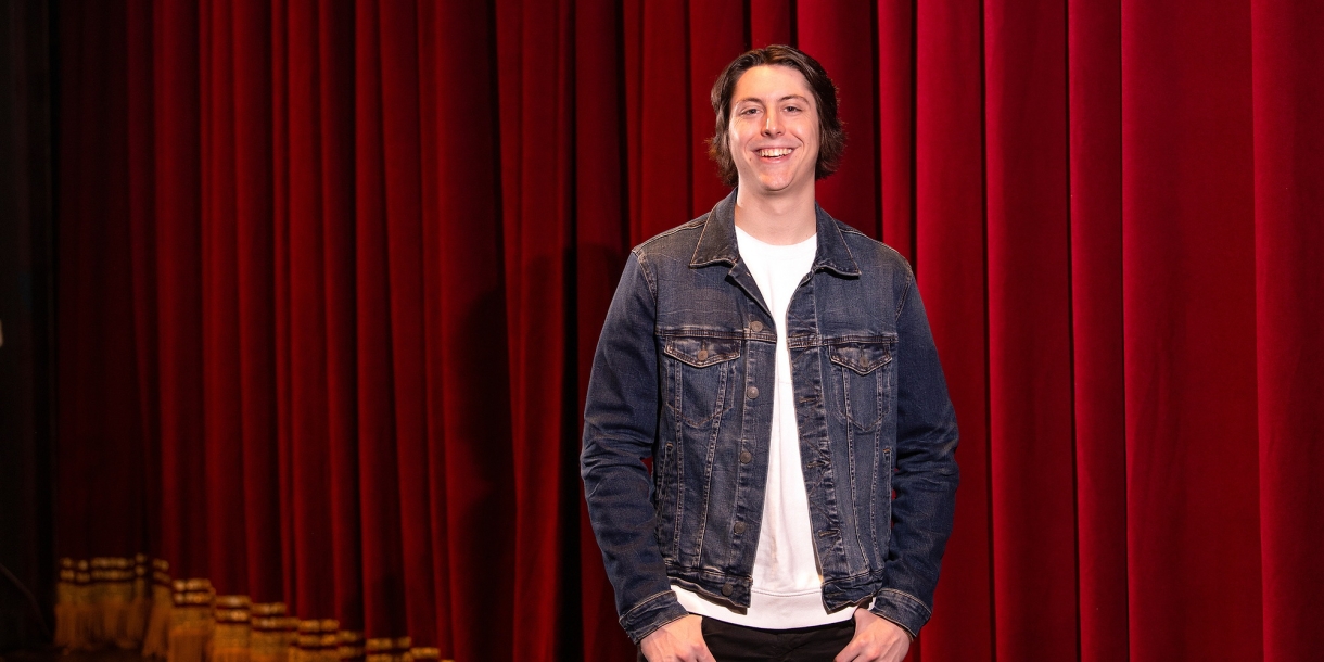 Reid Fournier stands on a stage, a red stage curtain is in the background.