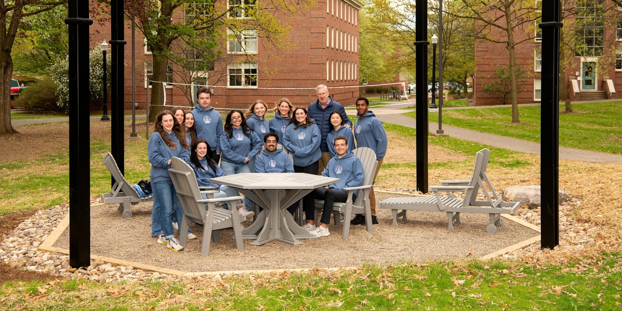 President John Bravman and Bucknell Student Government members from the Class of 2023 gather at a table on an octantal-shaped pit, with four pillars and stringed lights overhead.