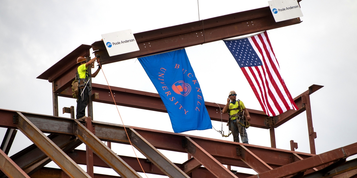 Two construction workers guide a beam into place on top of the building. American and Bucknell University flags hang from the beam.