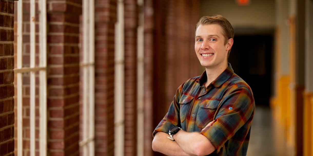 Chris Gregory wears a plaid shirt and stands in a hallway with window and brick trim.