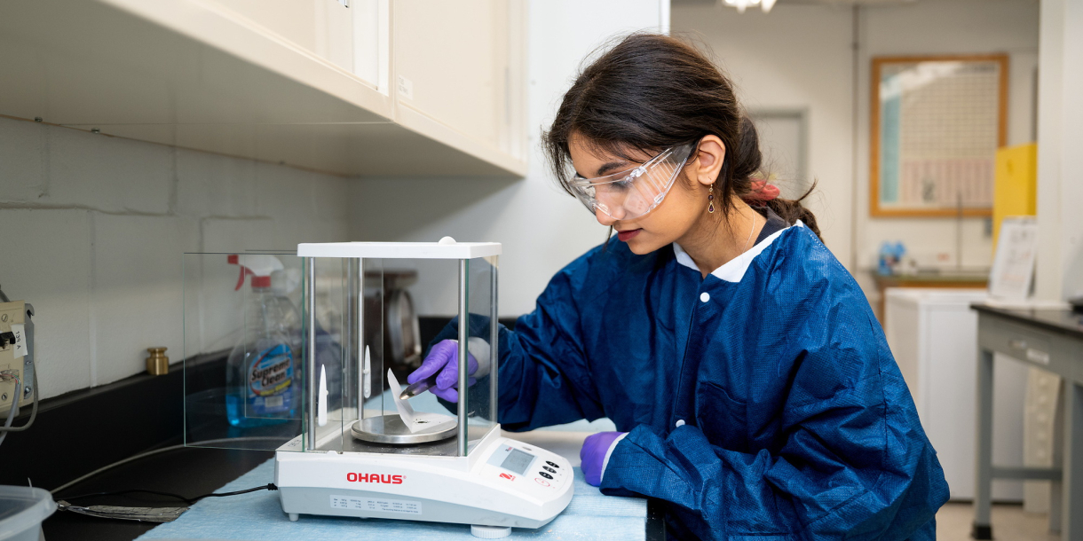 Priyanka Poplai '29 wears a blue lab coat, blue gloves, and safety goggles as she works in a lab space.