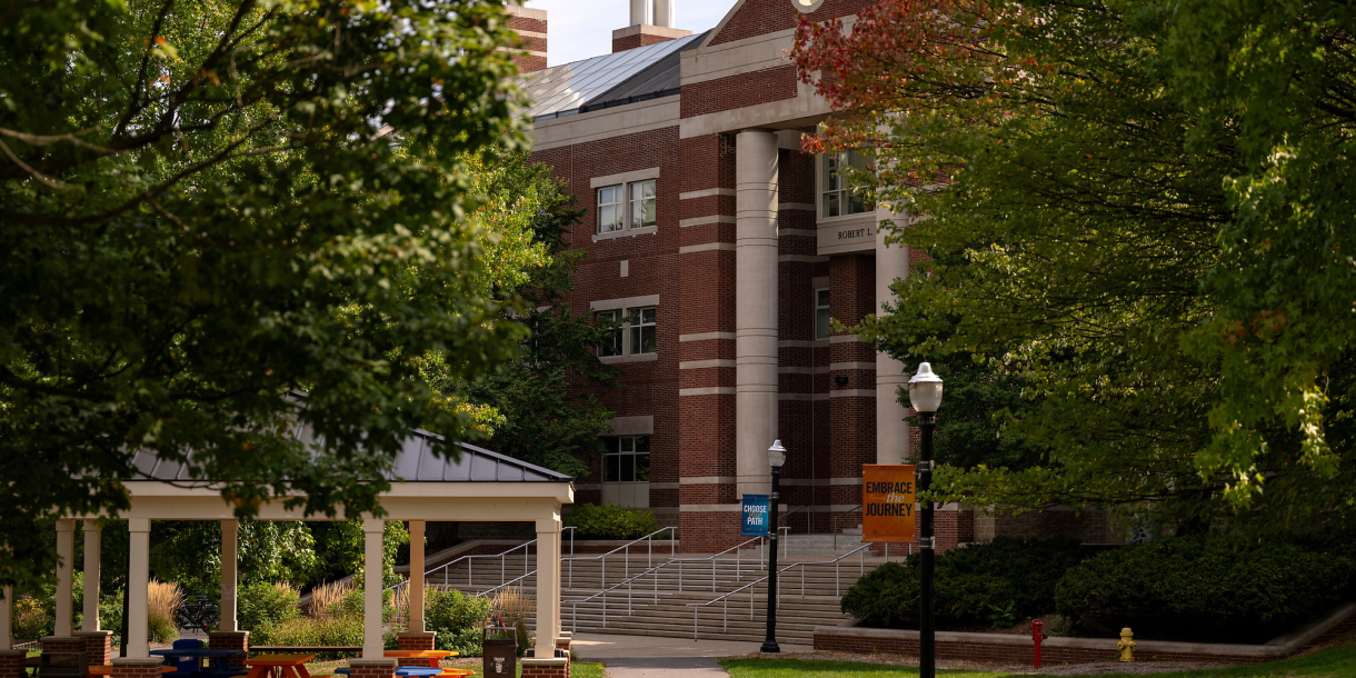 a scenic shot of campus features a covered patio with orange blue chairs, with Rooke Science Center in the backdrop and lush green foliage.