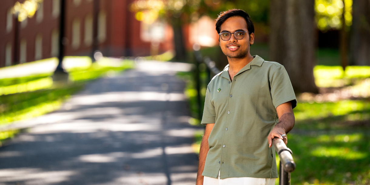 A Bucknell student wearing a green shirt stands outside on a sunny day with trees and a walking path in the background.