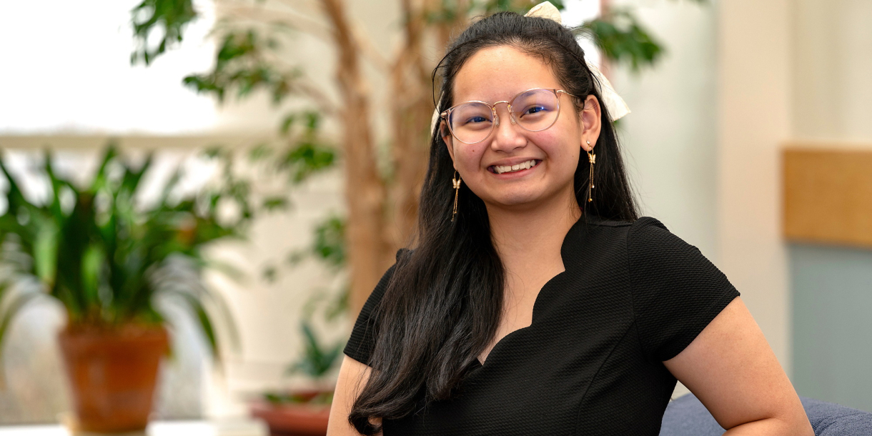 Angel Eunisse Yanga wears a black short-sleeved shirt and smiles in a room where there are potted plants in the background.
