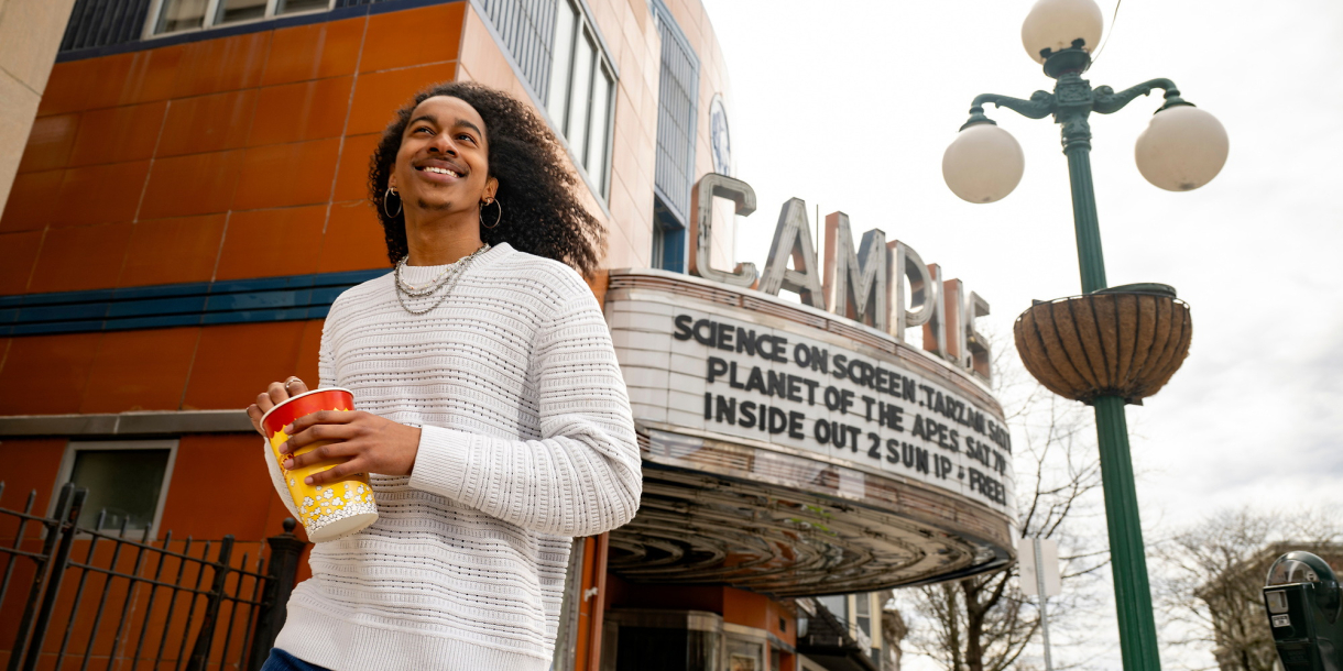 Caleb Warren smiles as he holds a small container of movie theatre popcorn; the marquee of the Campus Theatre is behind him.