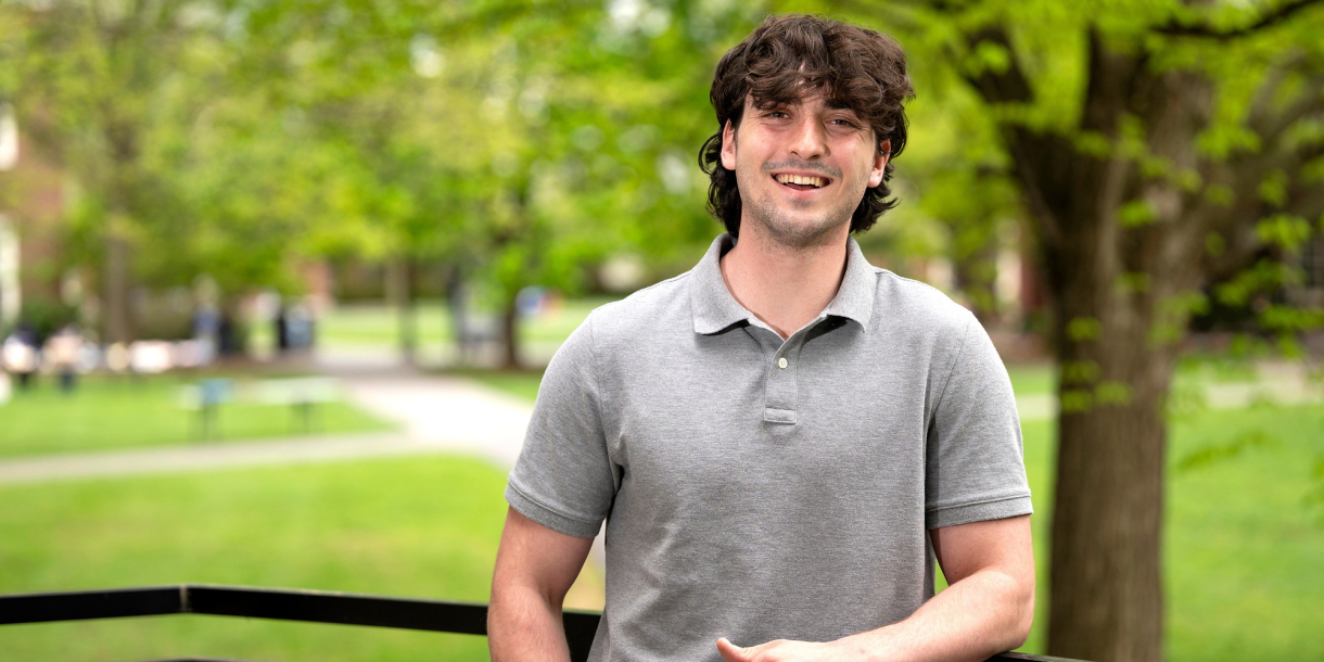 Michael Evanick stands outdoors on campus and smiles; green grass and trees are behind him.