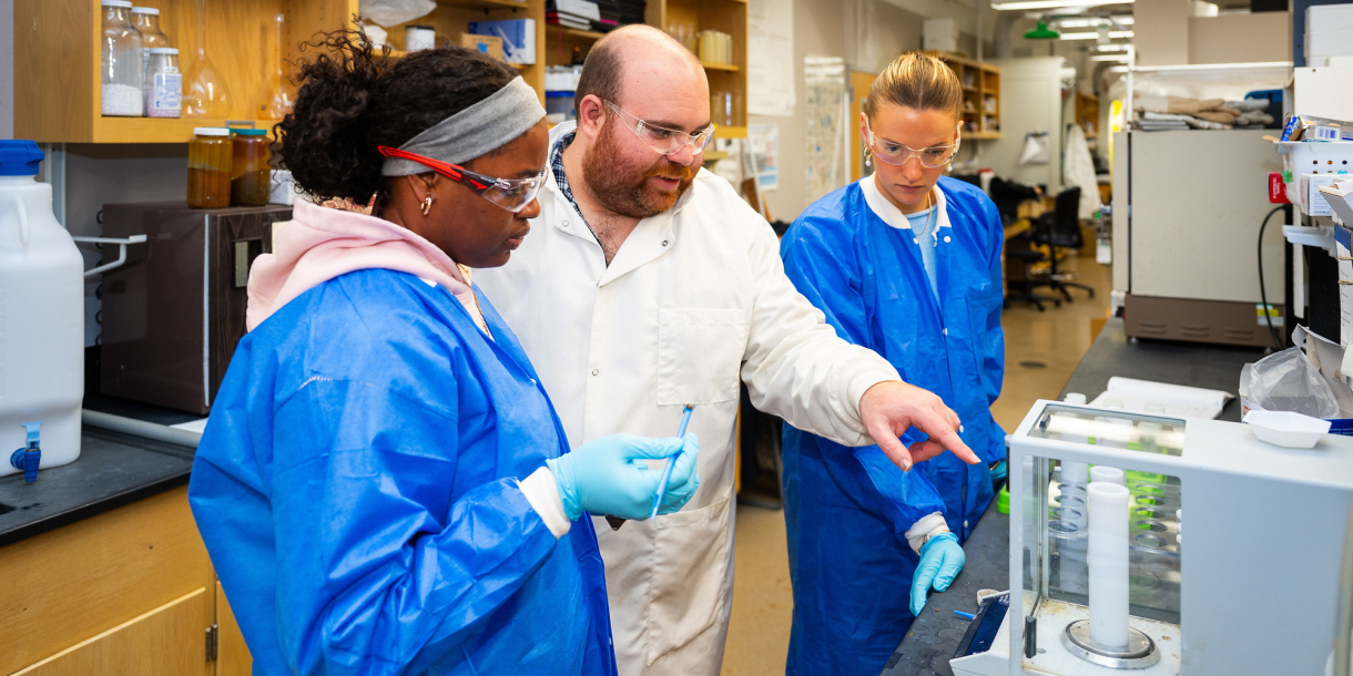 Two students wearing blue lab coats and a professor in a white lab coat stand inside of a laboratory.