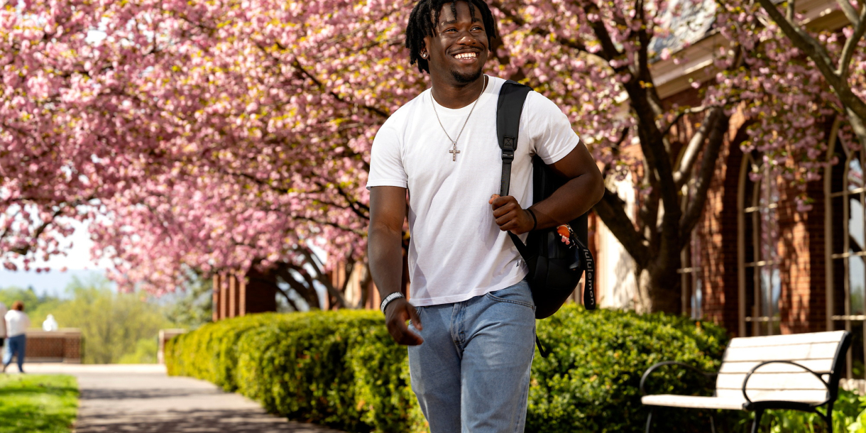 Momo Peters wears a white T-shirt and jeans with a backpack slung over his left shoulder as he smiles and walks on campus with pink cherry blossoms behind him.