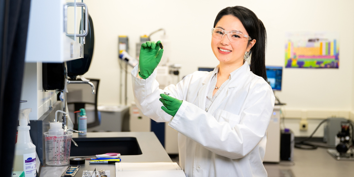 My Ly wears a white lab coat, safety goggles and green gloves while holding lab equipment in a lab setting.