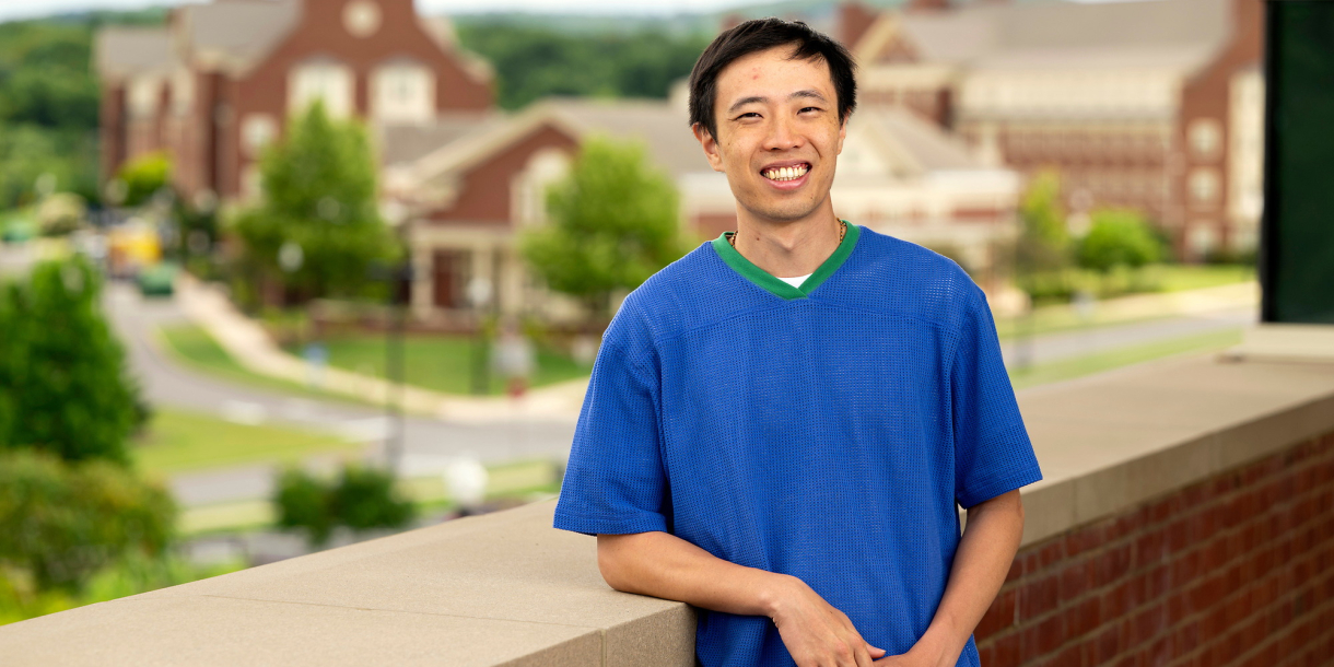 Sing Chun Lee wears a bright blue shirt and leans against a brick wall while smiling on campus