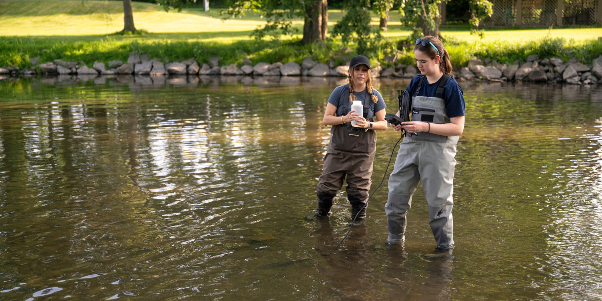 Two researchers wearing chest waders stand in a shallow waterway, one holding a sample bottle and the other checking a handheld device.
