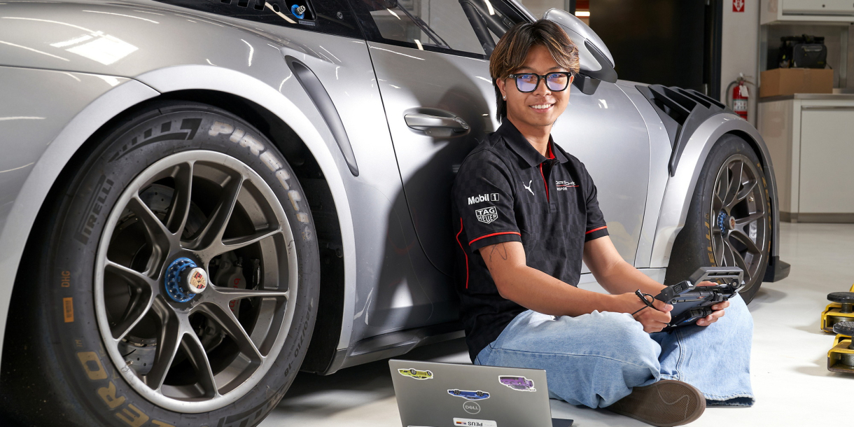Zach Ebiane sits on the ground with a laptop while leaning against a silver Porsche.