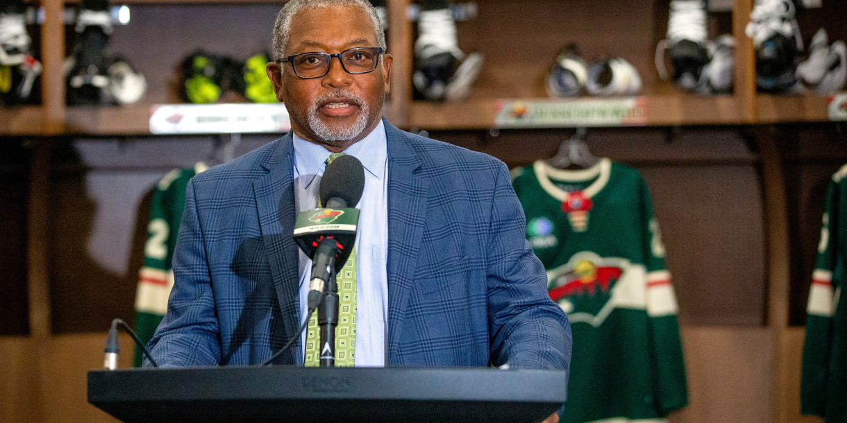 Joel Boyd stands at a podium and talks, with Minnesota Wild jerseys hanging behind him.