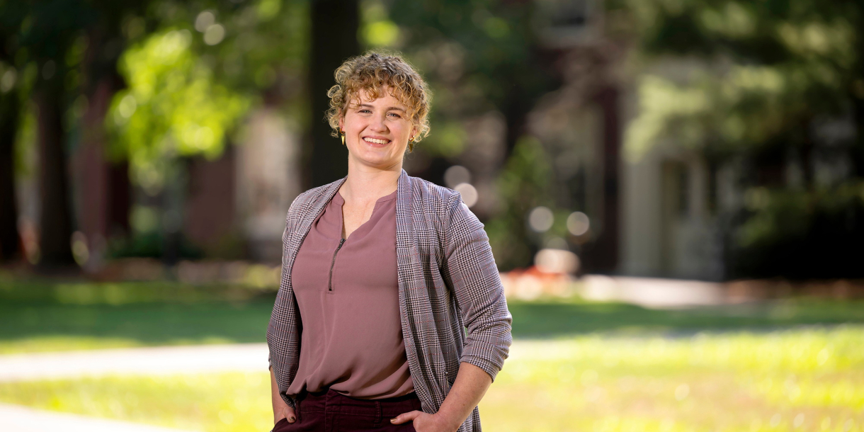 Annie Ross smiles with her hands in her pockets with greenery in the background.